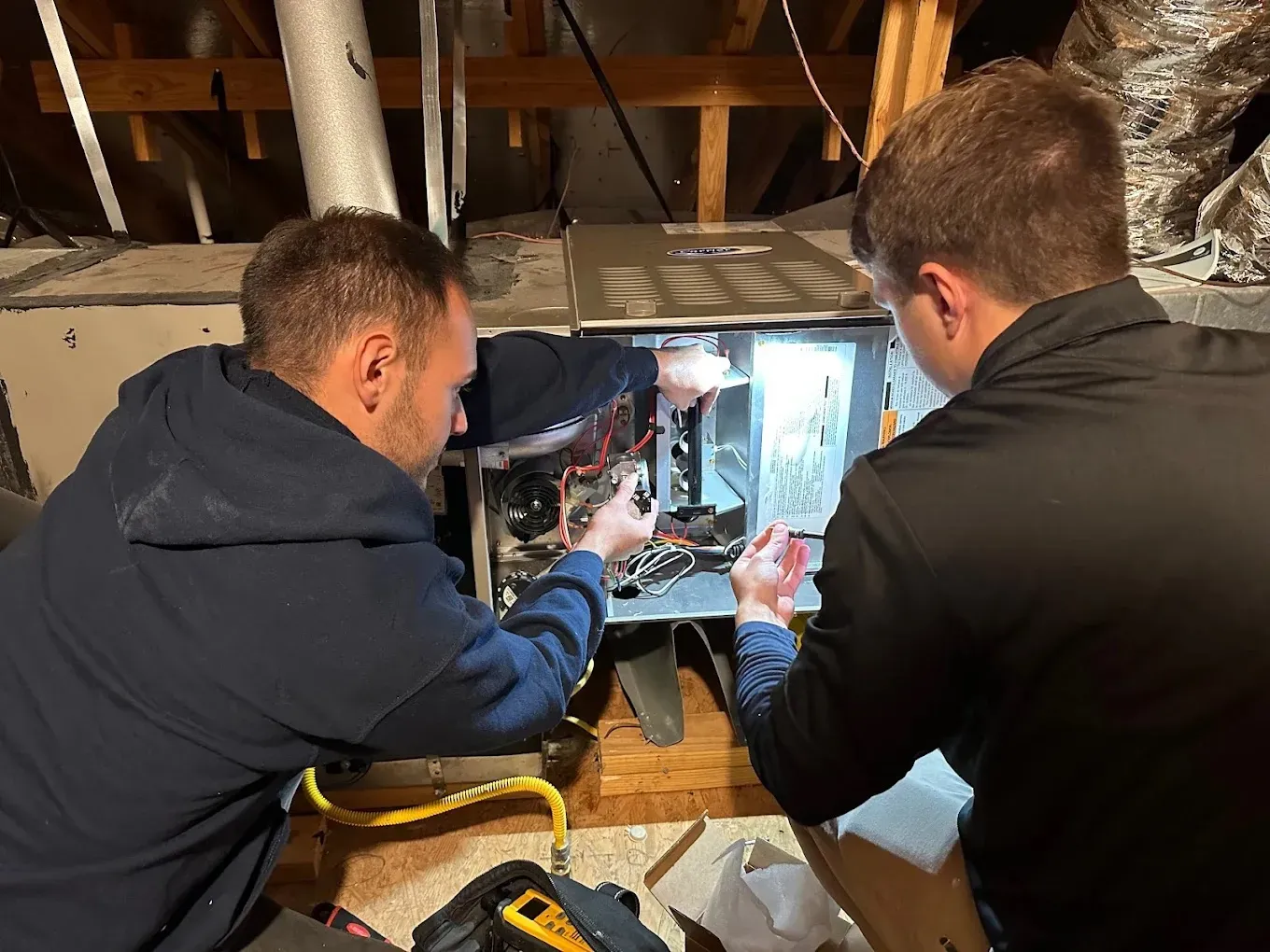 Two technicians repairing an HVAC unit in an attic space.